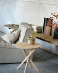 A minimalist living space featuring a light gray sofa, an Ethnicraft Mikado Side Table - Oak with a potted plant, and a rustic wooden shelf displaying books, a bowl, and autumn leaves in a vase against a textured white wall.