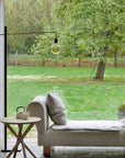 A cozy indoor nook features a cushioned daybed and an Ethnicraft Mikado Side Table - Oak with a teapot and cup. A large window reveals views of a lush green garden with trees and grass outside.