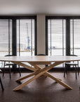 A modern meeting room with an Ethnicraft Mikado Meeting Table - Oak, featuring a distinctive intersecting base, surrounded by six black chairs and tall windows with horizontal blinds letting in natural light.