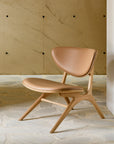 The Ethnicraft Eye Lounge Chair - Oak with Leather sits on a stone floor against a beige wall with visible dark bolts.