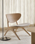 A minimalist living space featuring the Ethnicraft Eye Lounge Chair in oak with off-white fabric, a black floor lamp, a round wooden coffee table, and beige curtains. The room showcases neutral tones and modern, Scandinavian-style decor.