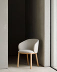 The Ethnicraft Unda Dining Chair, featuring light wooden legs and a light beige upholstered shell, sits by a large window against gray concrete walls.
