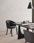 A modern minimalist office with a black desk, two dark gray Ethnicraft Unda Dining Chairs featuring upholstered shells, a geometric black pendant light, and workspace essentials on the desk, set against a textured light wall.