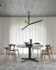 A modern dining space with a square black table, four Ethnicraft Bok Dining Chairs in natural teak, a black geometric ceiling light, sheer white curtains, and a stone-patterned floor for a minimalist, bright look.