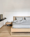 Minimalist bedroom with light wood floors, a solid European oak bed, gray bedding, and matching pillows. The Ethnicraft Nordic II Bedside Table holds a glass of water, books, and a tray; above hangs a black floating shelf with decor.