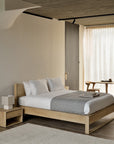 A minimalist bedroom with an Ethnicraft Nordic wooden bed in solid oak, white bedding, and a gray throw. The Ethnicraft Nordic II Bedside Table with a lamp is on the left; a chair and table sit beside sheer curtains streaming natural light.