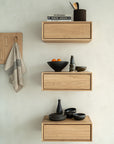 Three wooden floating shelves on a light wall display stacked books, a mortar and pestle, oranges in a bowl, dishes, and bottles. To the left, a striped towel hangs on the Ethnicraft Utilitile Wall Hanger.