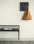 A minimalist interior features a black bench with a magazine, a brown bag hanging on the Ethnicraft Utilitile Wall Hanger against a light wall, and a textured rug partially covering the floor.