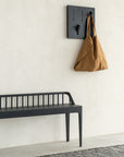 A minimalist scene features a black bench by a light textured wall. Above it, the Ethnicraft Utilitile Wall Hanger holds a large brown tote bag, while a gray woven rug adds texture to the floor.