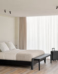 A minimalist bedroom features a sleek black bed frame, white bedding, beige blanket, and the Ethnicraft Bok Bench in Black Oak at the foot of the bed. A desk, chair, and suitcase sit near a large window with sheer white curtains.