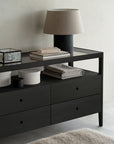 The Spindle Dresser - Black by Ethnicraft displays drawers, a beige table lamp, stacked books, decorative boxes, and neatly folded gray towels arranged on and beneath it against a light wall.
