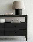 The Spindle Dresser - Black by Ethnicraft, featuring two drawers, displays a folded gray towel, round white box, stacked books, and a lamp with a black base and beige shade against a light-colored wall.