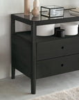 The Ethnicraft Spindle Dresser - Black is a modern, black-stained solid oak piece featuring two drawers and decorative items on top, set against a light-colored wall.