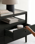 A person opens a black-stained Spindle Dresser by Ethnicraft, revealing folded linens and a round box inside. On top, stacked books, neatly folded fabric, and a black-base lamp complete the elegant display.