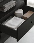 An open Ethnicraft Spindle Dresser - Black reveals neatly folded white and gray towels, a round white container, and a rectangular tan box inside, with more folded towels arranged on the shelf above.