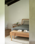 A minimalist bedroom with Ethnicraft’s Spindle Bed in oak, gray bedding, a matching bench, a nightstand with a plant and books, all set against neutral walls and European oak flooring.