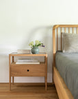 An Ethnicraft Spindle Bed - Oak with gray bedding stands against a white wall, beside a matching wooden nightstand that holds stacked books and a blue vase of white flowers.