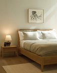 A minimalist bedroom features the Ethnicraft Spindle Bed - Oak, white and beige bedding, a lamp-topped bedside table, and framed abstract art on a pale wall. Soft natural light gently brightens the space.