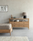 A minimalist bedroom featuring Ethnicraft furniture—a wooden bed, Spindle Dresser - Oak by Ethnicraft with four drawers, a gray lamp, decorative vases, stacked books, framed wall art, and neutral-toned decor.