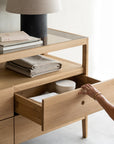 A person opens a drawer in the Ethnicraft Spindle Dresser - Oak, revealing neatly folded linens and a round container. The glass top displays stacked books, a lamp, and more folded fabric on the lower shelf.