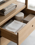 An open drawer of the Ethnicraft Spindle Dresser - Oak holds neatly folded towels, a round white container, and a rectangular beige box; additional towels are stacked on the shelf above.