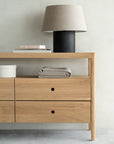 The Ethnicraft Spindle Dresser - Oak features four drawers and is styled with folded towels, a cylindrical box, books, and a beige-shaded table lamp on top, set against a light-colored wall.