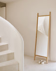 A minimalist interior features a curved white staircase, the Ethnicraft PI Floor Mirror in oak reflecting the stairs, and a pair of white sneakers neatly placed on the polished imperfect floor before the mirror.