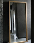 The Ethnicraft Light Frame Floor Mirror - Oak stands against a gray concrete wall in a minimalist room, reflecting a hallway and closed door.