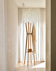 The Ethnicraft PI Coat Stand - Oak holds a beige garment and small brown bag in a bright room, positioned before sheer white floor-to-ceiling curtains that let natural light stream in.