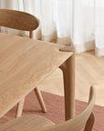 A close-up of the Ethnicraft PI Dining Table - Oak with two matching curved-back chairs sits on a pink rug over a herringbone wood floor near a window dressed in sheer white curtains.