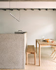 Minimalist kitchen with light wood furniture, a terrazzo island, Ethnicraft Pi Bench - Oak, table set with a white bowl of oranges and a glass of water, tiled flooring, exposed beam ceiling, and modern lighting.