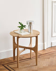 The Ethnicraft Pi Side Table - Oak features a small, lightweight solid oak build with curved legs. Shown on a wood floor, it holds a book, plant in a glass jar, and modern silver lamp against white walls and trim.