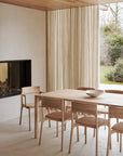 A minimalist dining room featuring Ethnicrafts PI Dining Chair in oak, a matching wooden table, beige curtains, built-in fireplace, and large windows with garden views. Natural light creates a calm, serene atmosphere.