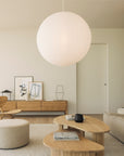A minimalist living room in neutral tones features a large round paper lantern and light wood furniture, including the Ethnicraft PI TV Cupboard crafted from solid oak. A low sofa faces framed art displayed on a sideboard, with a round ottoman placed on a rug. Sunlight pours in through a large window on the left.