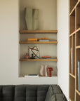 A modern living room features an Ethnicraft Wall Shelf - Oak displaying books, abstract vases, and décor above a gray tufted sofa. Soft beige, brown, and muted accent tones create a cozy atmosphere with a stylish neutral palette.