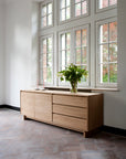 The Ethnicraft Wave Sideboard with three drawers and two cabinets sits beneath large windows, topped with two pink candles in holders and a glass vase of green foliage. Sunlight fills the minimalist space.