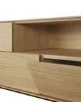 A close-up of the Ethnicraft Whitebird TV Cupboard showcases its solid oak build, open shelf, and smooth angular lines. The light wood finish emphasizes the natural grain and minimalist design.