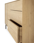 The Ethnicraft Whitebird Sideboard - 71 is a modern oak piece with a natural finish, clean lines, and black legs. A partially open large front drawer reveals its spacious storage.