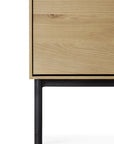 Close-up of the bottom corner of the Ethnicraft Whitebird Sideboard - 71 in solid oak, featuring a light natural finish and a black cylindrical metal leg, set against a white background.