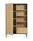 The Ethnicraft Blackbird Storage Cupboard is crafted in European oak with a black frame and tall legs. It offers shelves and a partially open door on the left, plus open shelves and a closed drawer on the right.