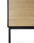 Close-up of the lower corner of the Ethnicraft Blackbird Sideboard – 2 doors + 3 drawers, crafted in European oak with a natural finish, highlighting a black frame and single cylindrical black leg.