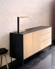 The Blackbird Sideboard - 2 doors + 3 drawers by Ethnicraft, with black metal sides and European oak drawers, stands against a white brick wall, complemented by a minimalist lamp and wooden stool on a dark tiled floor.