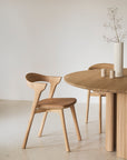 A minimalist dining area features a light wooden chair next to Ethnicrafts Roller Max Round Dining Table in oak, topped with a tall white vase holding delicate branches and two small cylinders, set against plain light walls and flooring.