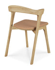 The Ethnicraft Bok Dining Chair features solid oak construction, angled legs, a curved backrest, and a Sienna leather seat. Shown from the rear side angle against a white background.