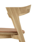 A close-up side view of the Ethnicraft Bok Dining Chair in solid oak with a sienna leather seat, showcasing its curved backrest, minimalist design, and elegant European oak wood grain.