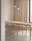 A minimalist dining area showcases the Ethnicraft Bok Extendable Dining Table - Oak with matching chairs on a speckled floor, tall beige curtains, and two modern pendant lights with exposed bulbs above.