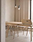 A minimalist dining room features light wooden chairs and the Bok Extendable Dining Table - Oak by Ethnicraft, one section open to reveal storage. Beige curtains cover a large window, with two modern pendant lights overhead.