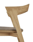 Side view of the Bok Dining Chair - Oak with Warm Grey Fabric by Ethnicraft, showcasing angled solid oak legs, a curved backrest, and a cushioned warm grey seat. This modern chair is set against a white background.