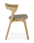 The Bok Dining Chair by Ethnicraft features solid oak, angled legs, a curved backrest, and a warm grey cushioned seat. Shown from the side on a white background.