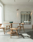 A bright, minimalist cafe with natural light features Ethnicraft’s Torsion Dining Table - Oak on a concrete floor, paired with chairs, a large window, white brick wall, and a mirror reflecting vertical striped decor.